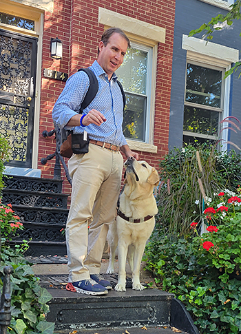 In front of Pat's red brick house, Hogan and Pat stand on a landing of black cast iron steps. Hogan is focused on the hotdog treat in Pat's right hand. Pat is wearing a patterned blue shirt, khakis, and a black backpack. Streaks of morning sun filters through a nearby tree and red flowers surround the steps. Hogan's training clicker can be seen on Pat's right wrist.