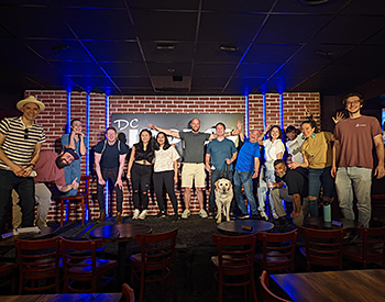 Pat and Hogan pose with their classmates on the main DC improv Comedy Club stage. There are 13 students standing with Chris, the instructor, and Hogan. The group is in front of a brick wall with the black letters of the Club's sign. Blue LED lights can be seen, along with bright stage lights. One student gives a thumbs up, while others gesture playfully. Pat wears a teal-colored T-shirt and dark jeans.