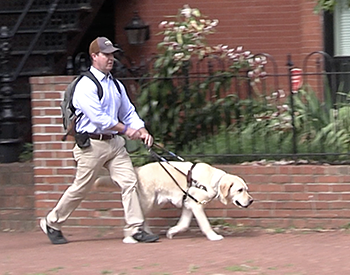 Team Hogan walks quickly down the sidewalk. Pat has on a black backpack and blue dress shirt, sleeves rolled halfway up his forearm. Hogan's head is slightly down as he focuses on guiding. A classic brick Capitol Hill house with an iron fence can be seen behind the two.